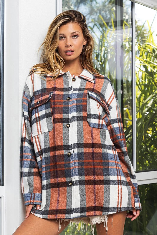 A woman in a Textured Shirt With Big Checkered Point leans casually against a wall, smiling while looking to the side. There are plants and a window in the background.