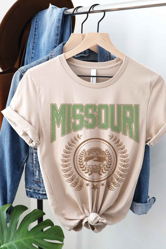 A person wearing a 100% cotton MISSOURI GRAPHIC TEE in white, paired with light blue jeans and a beige jacket draped over their shoulders.