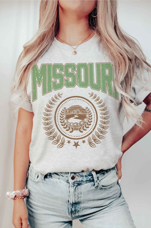 A person wearing a 100% cotton MISSOURI GRAPHIC TEE in white, paired with light blue jeans and a beige jacket draped over their shoulders.