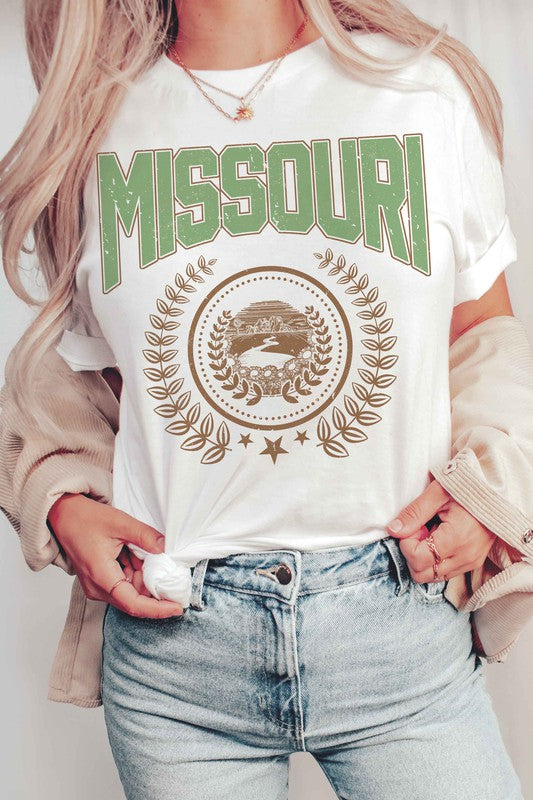 A person wearing a 100% cotton MISSOURI GRAPHIC TEE in white, paired with light blue jeans and a beige jacket draped over their shoulders.