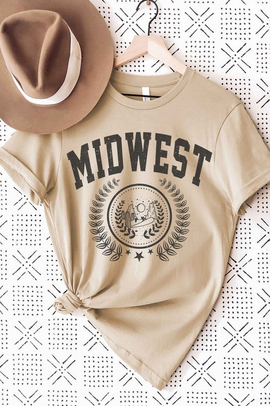 A person wearing the MIDWEST GRAPHIC TEE, a white t-shirt made of 100% cotton, featuring "MIDWEST" printed in large letters above a graphic of two people, possibly astronauts. The background includes a sun and trees, surrounded by leaves. This tee offers unisex sizing for a perfect fit.