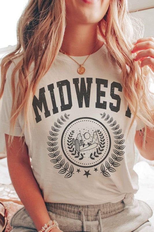 A person wearing the MIDWEST GRAPHIC TEE, a white t-shirt made of 100% cotton, featuring "MIDWEST" printed in large letters above a graphic of two people, possibly astronauts. The background includes a sun and trees, surrounded by leaves. This tee offers unisex sizing for a perfect fit.