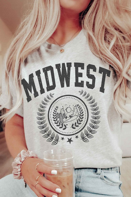 A person wearing the MIDWEST GRAPHIC TEE, a white t-shirt made of 100% cotton, featuring "MIDWEST" printed in large letters above a graphic of two people, possibly astronauts. The background includes a sun and trees, surrounded by leaves. This tee offers unisex sizing for a perfect fit.