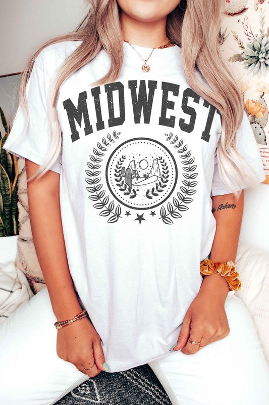 A person wearing the MIDWEST GRAPHIC TEE, a white t-shirt made of 100% cotton, featuring "MIDWEST" printed in large letters above a graphic of two people, possibly astronauts. The background includes a sun and trees, surrounded by leaves. This tee offers unisex sizing for a perfect fit.