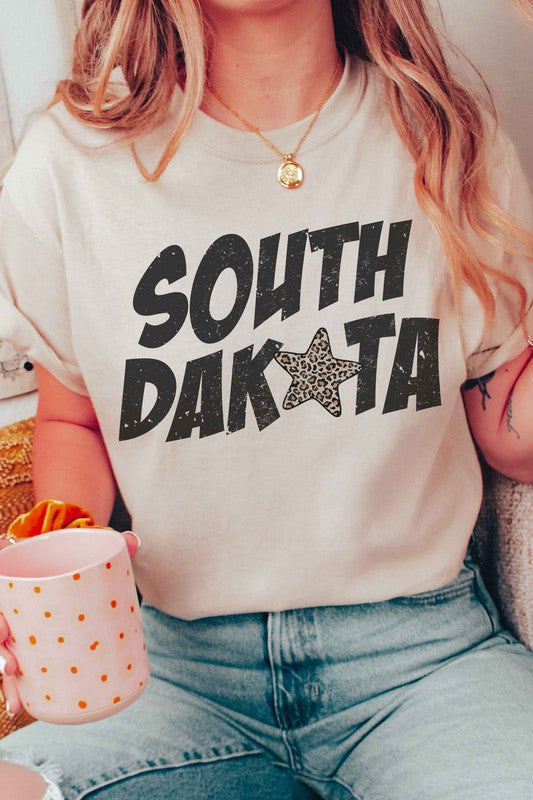 A person is wearing a SOUTH DAKOTA LEOPARD STAR GRAPHIC TEE in beige with unisex sizing, and holding a pink polka dot mug filled with orange slices.