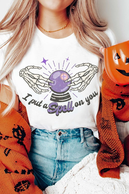 Person wearing the I PUT A SPELL ON YOU GRAPHIC TEE, featuring skeletal hands around a crystal ball and the text "I PUT A SPELL ON YOU." Holding a white mug with orange polka dots. Unisex sizing for a perfect fit.