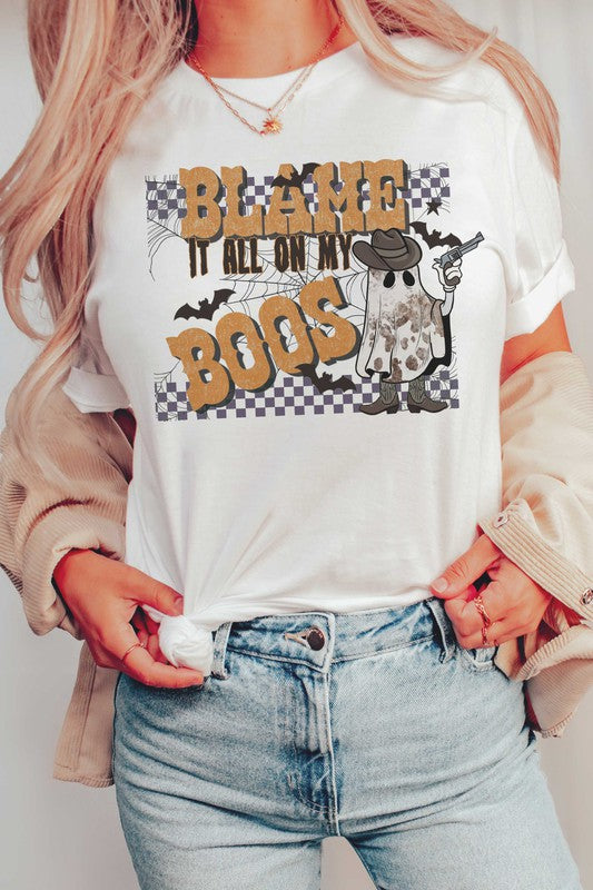 A person wearing the BLAME IT ALL ON MY BOOS GRAPHIC TEE, made from 100% cotton and featuring a cowboy ghost beside the text "Blame it all on my boos," holds a mug while showcasing their wrist adorned with bracelets. The unisex sizing ensures everyone can join in on the spooky fun.