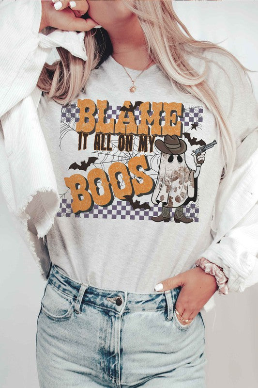 A person wearing the BLAME IT ALL ON MY BOOS GRAPHIC TEE, made from 100% cotton and featuring a cowboy ghost beside the text "Blame it all on my boos," holds a mug while showcasing their wrist adorned with bracelets. The unisex sizing ensures everyone can join in on the spooky fun.