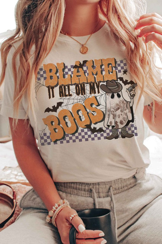 A person wearing the BLAME IT ALL ON MY BOOS GRAPHIC TEE, made from 100% cotton and featuring a cowboy ghost beside the text "Blame it all on my boos," holds a mug while showcasing their wrist adorned with bracelets. The unisex sizing ensures everyone can join in on the spooky fun.