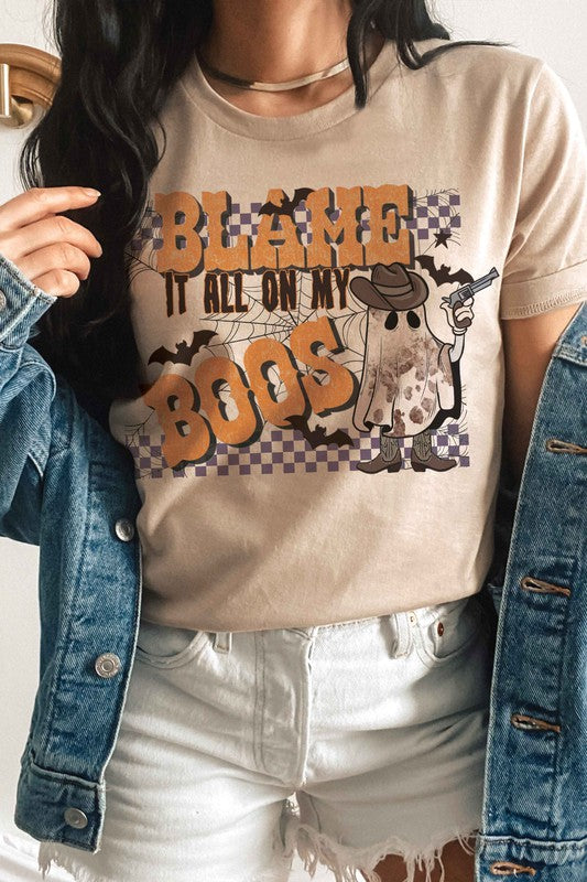 A person wearing the BLAME IT ALL ON MY BOOS GRAPHIC TEE, made from 100% cotton and featuring a cowboy ghost beside the text "Blame it all on my boos," holds a mug while showcasing their wrist adorned with bracelets. The unisex sizing ensures everyone can join in on the spooky fun.