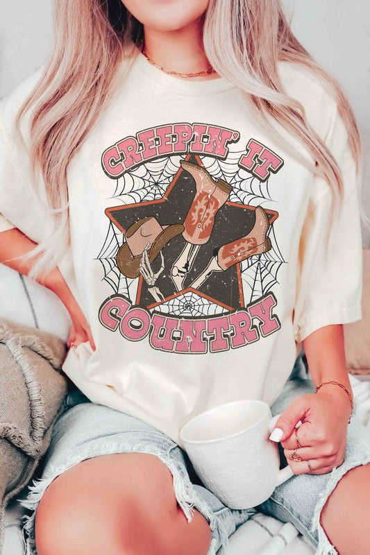 A person wearing the CREEPIN' IT COUNTRY GRAPHIC TEE, which features a design of cowboy boots, a hat, and spiderwebs, paired with a necklace. This stylish t-shirt comes in unisex sizing.