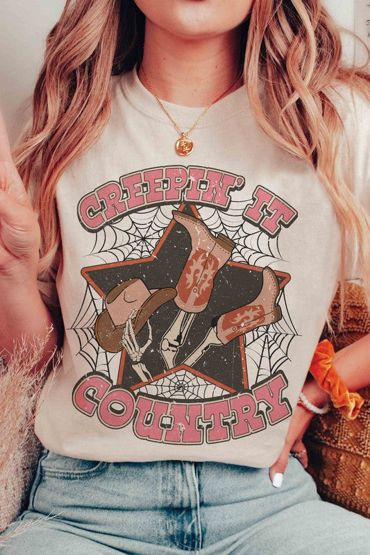 A person wearing the CREEPIN' IT COUNTRY GRAPHIC TEE, which features a design of cowboy boots, a hat, and spiderwebs, paired with a necklace. This stylish t-shirt comes in unisex sizing.