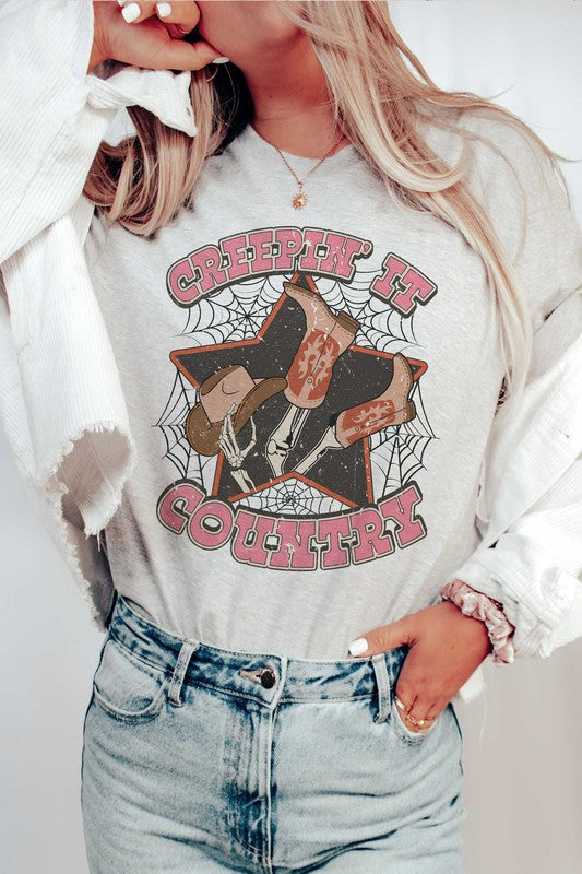 A person wearing the CREEPIN' IT COUNTRY GRAPHIC TEE, which features a design of cowboy boots, a hat, and spiderwebs, paired with a necklace. This stylish t-shirt comes in unisex sizing.