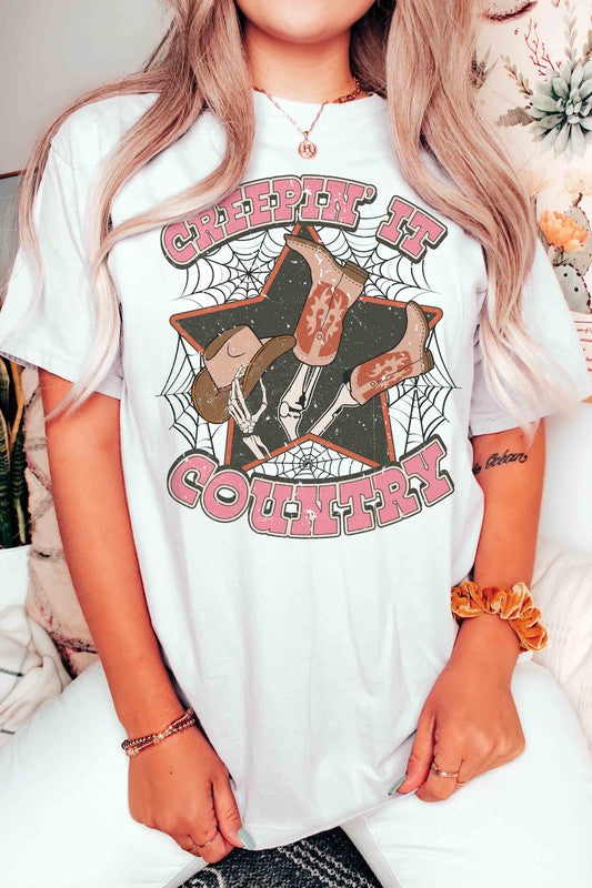 A person wearing the CREEPIN' IT COUNTRY GRAPHIC TEE, which features a design of cowboy boots, a hat, and spiderwebs, paired with a necklace. This stylish t-shirt comes in unisex sizing.