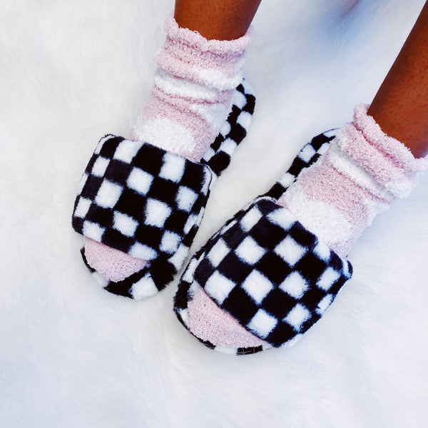 Four pairs of Luxe Lounge Checker Cozy Slippers in colors black, green, and tan are arranged on a white fluffy surface. Each pair boasts non-slip soles and a soft fabric construction for ultimate comfort.