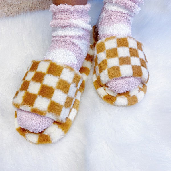 Four pairs of Luxe Lounge Checker Cozy Slippers in colors black, green, and tan are arranged on a white fluffy surface. Each pair boasts non-slip soles and a soft fabric construction for ultimate comfort.