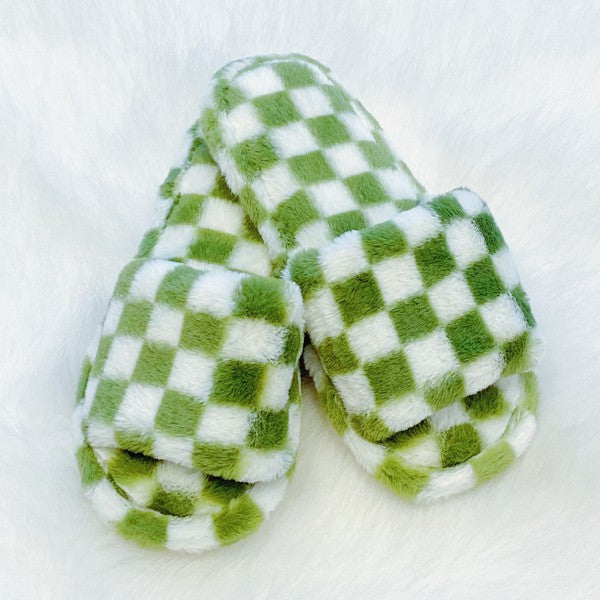 Four pairs of Luxe Lounge Checker Cozy Slippers in colors black, green, and tan are arranged on a white fluffy surface. Each pair boasts non-slip soles and a soft fabric construction for ultimate comfort.