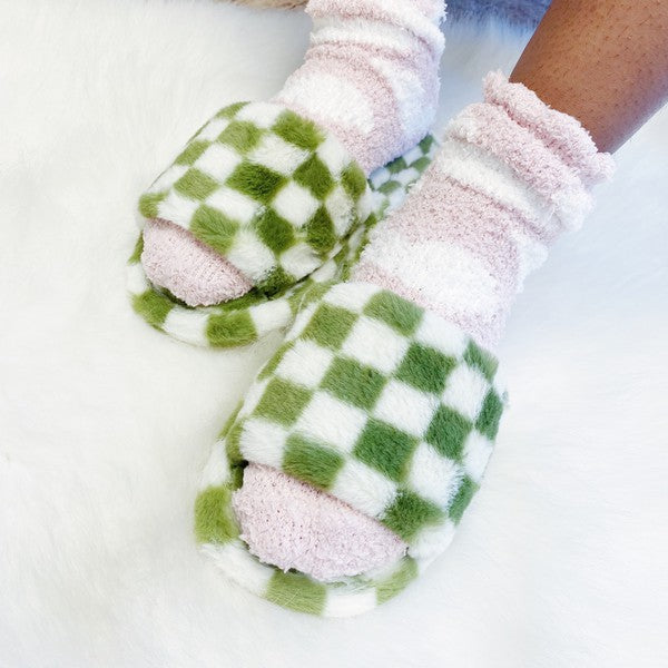 Four pairs of Luxe Lounge Checker Cozy Slippers in colors black, green, and tan are arranged on a white fluffy surface. Each pair boasts non-slip soles and a soft fabric construction for ultimate comfort.