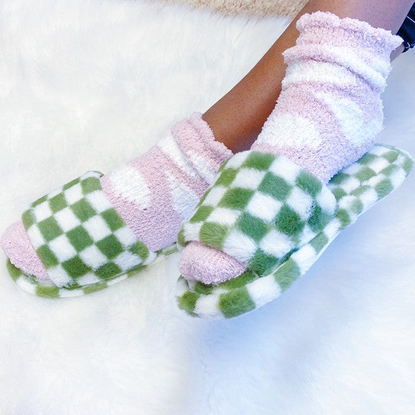 Four pairs of Luxe Lounge Checker Cozy Slippers in colors black, green, and tan are arranged on a white fluffy surface. Each pair boasts non-slip soles and a soft fabric construction for ultimate comfort.