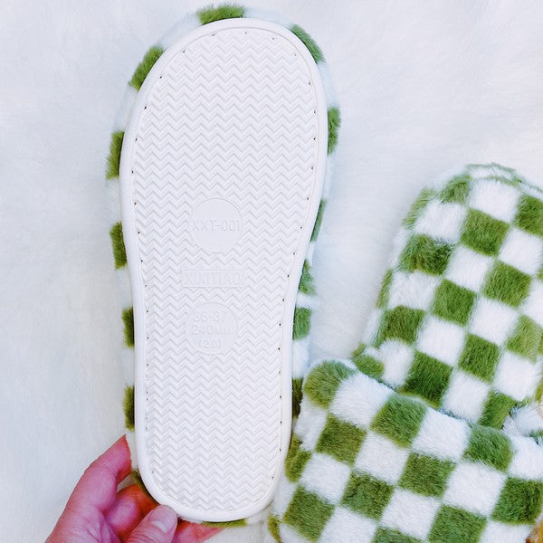 Four pairs of Luxe Lounge Checker Cozy Slippers in colors black, green, and tan are arranged on a white fluffy surface. Each pair boasts non-slip soles and a soft fabric construction for ultimate comfort.