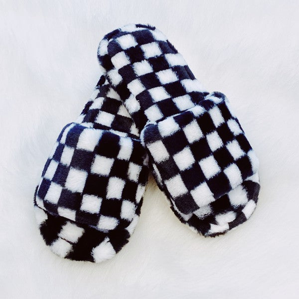 Four pairs of Luxe Lounge Checker Cozy Slippers in colors black, green, and tan are arranged on a white fluffy surface. Each pair boasts non-slip soles and a soft fabric construction for ultimate comfort.