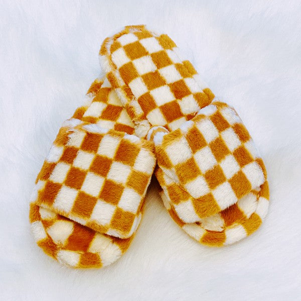 Four pairs of Luxe Lounge Checker Cozy Slippers in colors black, green, and tan are arranged on a white fluffy surface. Each pair boasts non-slip soles and a soft fabric construction for ultimate comfort.