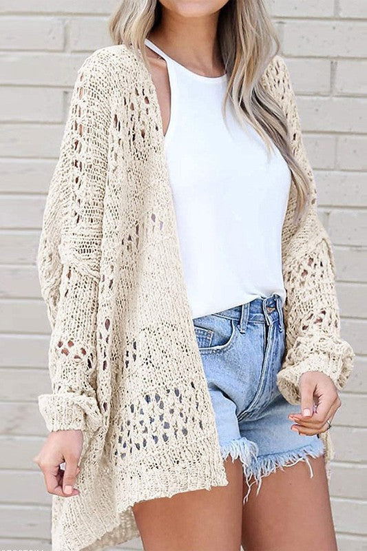 A person wearing a Crochet Dolman Knit Sleeve Cardigan in light beige, a white t-shirt, and blue denim shorts stands with their hands in their pockets.