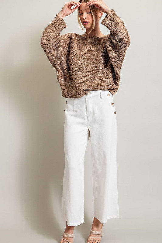 A woman smiles while wearing the Loose Fit Knit Top featuring a boat neckline, paired with white pants with button embellishments on the side. She stands against a plain background.