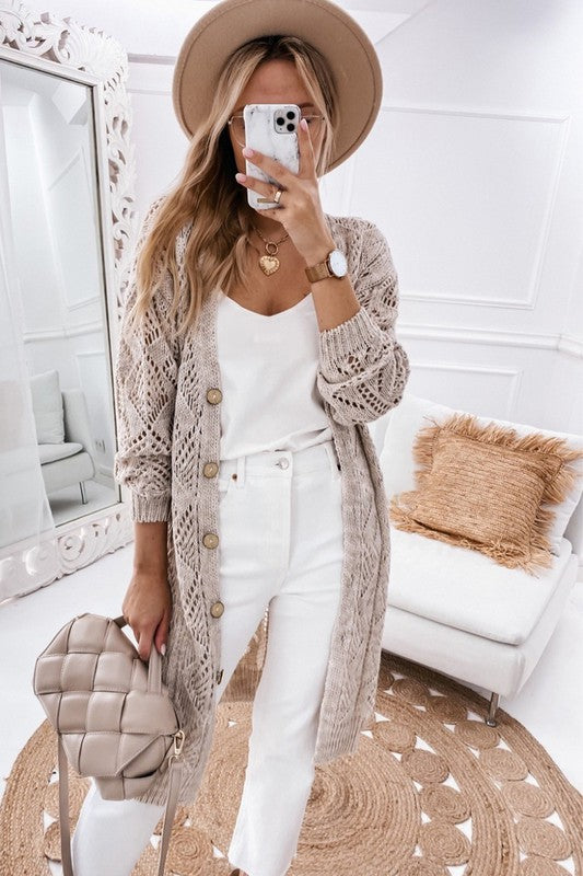 A person wearing a beige hat, white outfit, and an eyelet sweater button cream cardigan with a hollowed knit design takes a mirror selfie in a white, modern room with a beige purse.