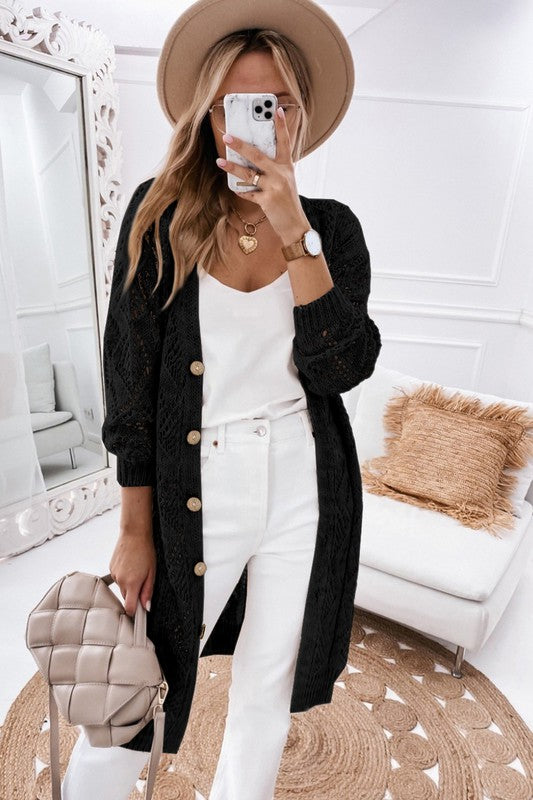 A person wearing a beige hat, white outfit, and an eyelet sweater button cream cardigan with a hollowed knit design takes a mirror selfie in a white, modern room with a beige purse.