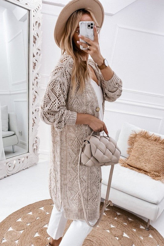 A person wearing a beige hat, white outfit, and an eyelet sweater button cream cardigan with a hollowed knit design takes a mirror selfie in a white, modern room with a beige purse.