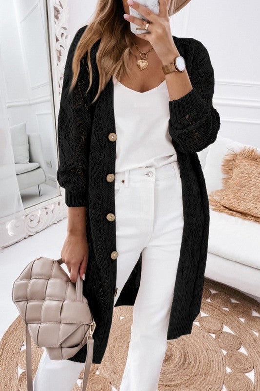 A person wearing a beige hat, white outfit, and an eyelet sweater button cream cardigan with a hollowed knit design takes a mirror selfie in a white, modern room with a beige purse.