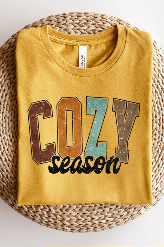 An "UNISEX SHORT SLEEVE" orange graphic tee hangs on a hanger, showcasing the words "COZY season" in a vibrant and bold font. This 100% cotton creation is perfect for anyone looking to embrace comfort and style.