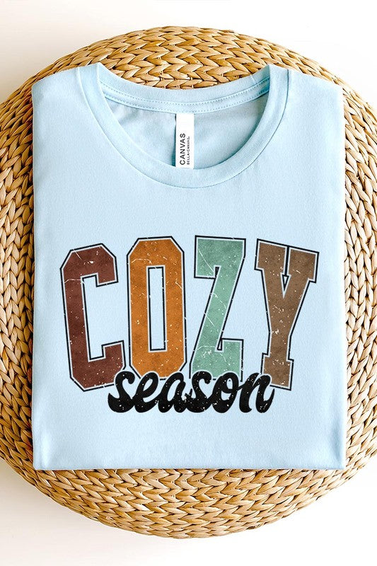 An "UNISEX SHORT SLEEVE" orange graphic tee hangs on a hanger, showcasing the words "COZY season" in a vibrant and bold font. This 100% cotton creation is perfect for anyone looking to embrace comfort and style.