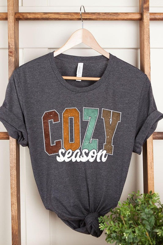 An "UNISEX SHORT SLEEVE" orange graphic tee hangs on a hanger, showcasing the words "COZY season" in a vibrant and bold font. This 100% cotton creation is perfect for anyone looking to embrace comfort and style.