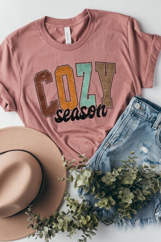 An "UNISEX SHORT SLEEVE" orange graphic tee hangs on a hanger, showcasing the words "COZY season" in a vibrant and bold font. This 100% cotton creation is perfect for anyone looking to embrace comfort and style.