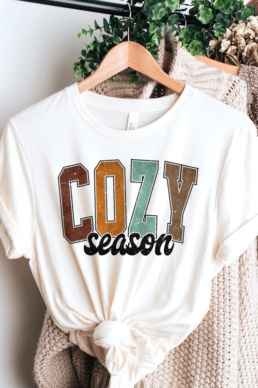 An "UNISEX SHORT SLEEVE" orange graphic tee hangs on a hanger, showcasing the words "COZY season" in a vibrant and bold font. This 100% cotton creation is perfect for anyone looking to embrace comfort and style.