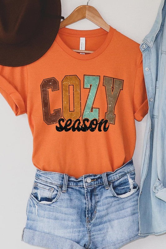 An "UNISEX SHORT SLEEVE" orange graphic tee hangs on a hanger, showcasing the words "COZY season" in a vibrant and bold font. This 100% cotton creation is perfect for anyone looking to embrace comfort and style.