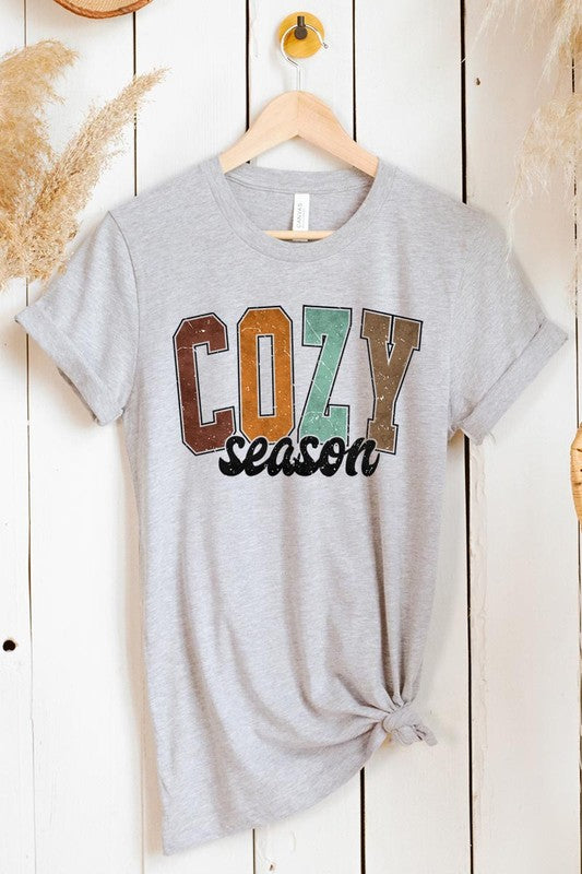 An "UNISEX SHORT SLEEVE" orange graphic tee hangs on a hanger, showcasing the words "COZY season" in a vibrant and bold font. This 100% cotton creation is perfect for anyone looking to embrace comfort and style.