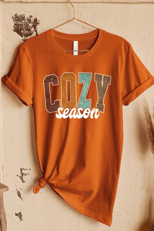 An "UNISEX SHORT SLEEVE" orange graphic tee hangs on a hanger, showcasing the words "COZY season" in a vibrant and bold font. This 100% cotton creation is perfect for anyone looking to embrace comfort and style.