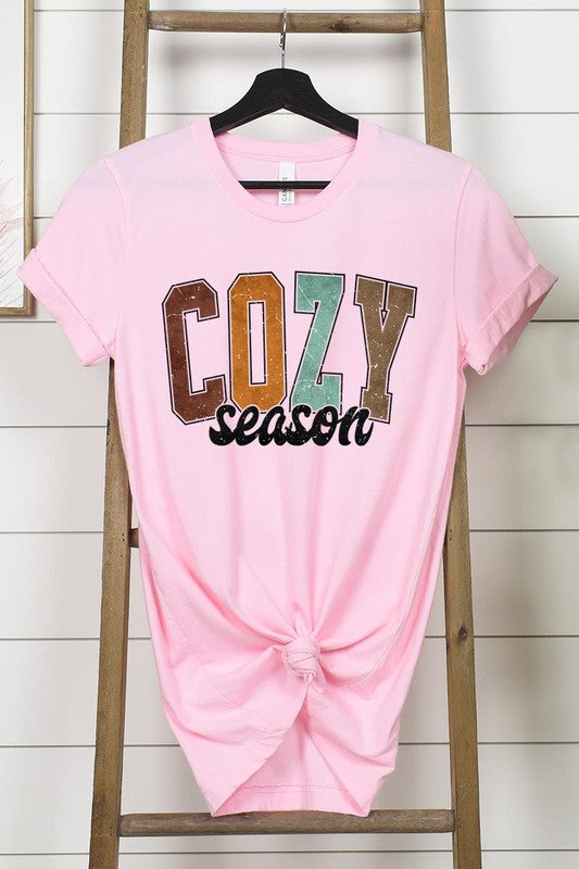 An "UNISEX SHORT SLEEVE" orange graphic tee hangs on a hanger, showcasing the words "COZY season" in a vibrant and bold font. This 100% cotton creation is perfect for anyone looking to embrace comfort and style.