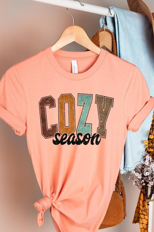 An "UNISEX SHORT SLEEVE" orange graphic tee hangs on a hanger, showcasing the words "COZY season" in a vibrant and bold font. This 100% cotton creation is perfect for anyone looking to embrace comfort and style.