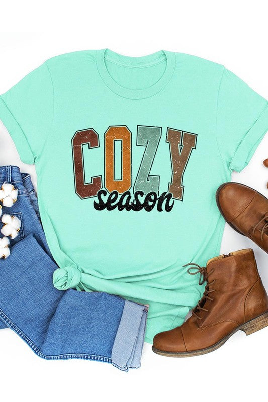 An "UNISEX SHORT SLEEVE" orange graphic tee hangs on a hanger, showcasing the words "COZY season" in a vibrant and bold font. This 100% cotton creation is perfect for anyone looking to embrace comfort and style.