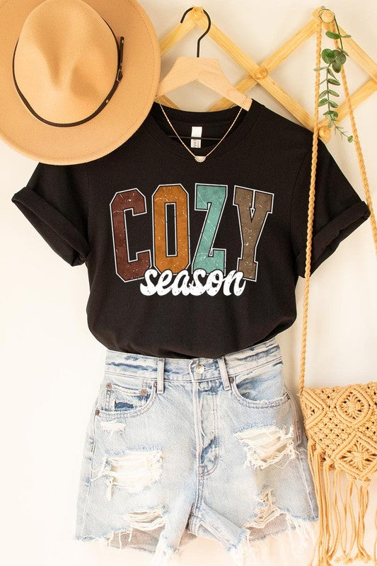 An "UNISEX SHORT SLEEVE" orange graphic tee hangs on a hanger, showcasing the words "COZY season" in a vibrant and bold font. This 100% cotton creation is perfect for anyone looking to embrace comfort and style.