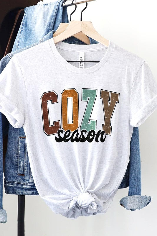 An "UNISEX SHORT SLEEVE" orange graphic tee hangs on a hanger, showcasing the words "COZY season" in a vibrant and bold font. This 100% cotton creation is perfect for anyone looking to embrace comfort and style.