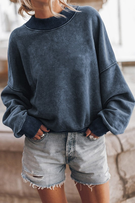 A person wearing a Mineral Washed Acid dye Sweatshirt Pullover with a drop shoulder design and distressed denim shorts stands with hands in pockets.
