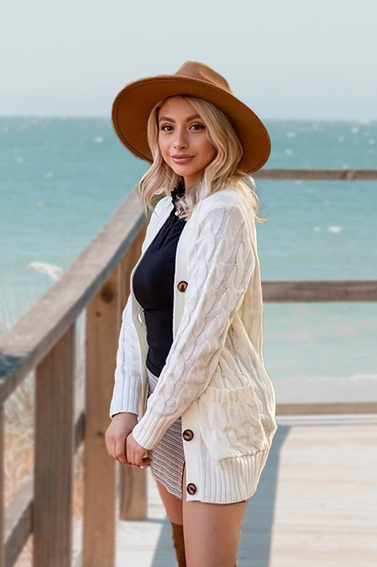 A woman wearing a beige hat, black top, white cable knit button cardigan with pocket, and a patterned polyester skirt stands on a wooden walkway by the sea.