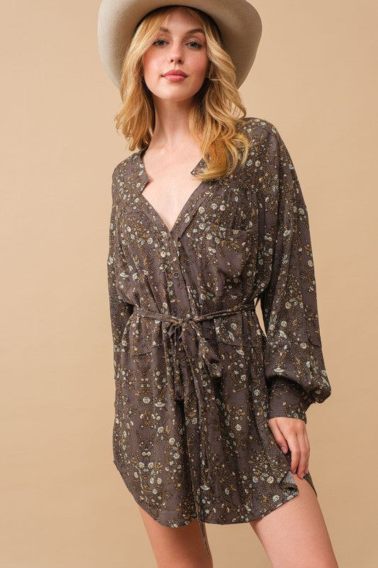 A person stands against a beige background, wearing a cowboy hat, a belt, cowboy boots, and the Rayon Crinkle Ditsy Floral Raw Shift Shirt Dress in gray.