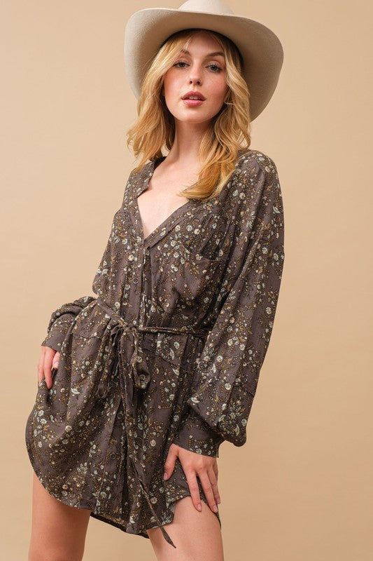 A person stands against a beige background, wearing a cowboy hat, a belt, cowboy boots, and the Rayon Crinkle Ditsy Floral Raw Shift Shirt Dress in gray.