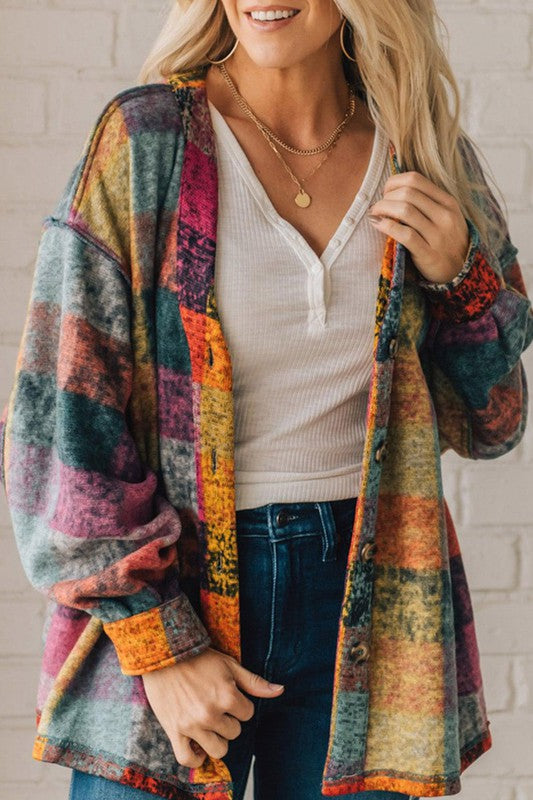 A person wearing a Mustard Red plaid button Shacket Cardigan Sweater made of 100% polyester over a white henley top and dark jeans, standing against a white brick wall.